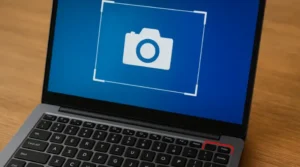Gambar menunjukkan laptop modern yang terbuka di atas meja kayu. Layar menampilkan antarmuka aplikasi kamera virtual dengan ikon kamera putih besar di tengah bingkai putar berwarna biru. Pada keyboard hitamnya, tombol "Prnt Scrn" (cetak layar) diberi garis merah tebal untuk menonjolkan fungsinya sebagai cara ss di komputer. Desain keseluruhan minimalis dan profesional, menggabungkan elemen teknologi dengan nuansa visual yang bersih.